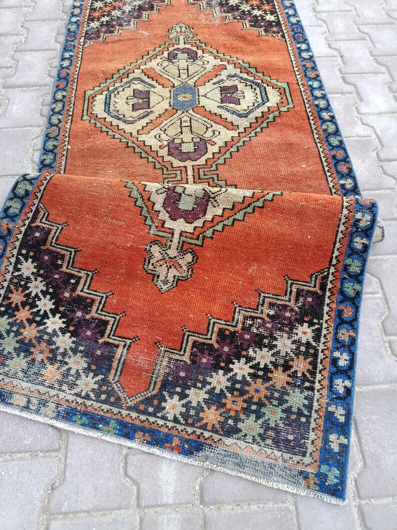 May include: An orange and blue vintage runner rug with geometric patterns. The rug features a central diamond design with cream, blue, and purple accents. The border is a dark blue with a repeating floral pattern. The rug is folded over.