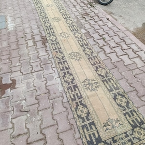 Può includere: Un tappeto lungo e stretto con un centro color crema e motivi floreali e geometrici ripetuti in grigio scuro. Il bordo presenta un complesso motivo grigio scuro di forme intrecciate ed elementi floreali, rifinito con un bordo rosso.