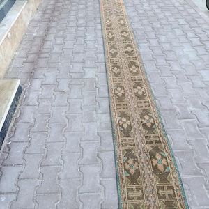May include: A long, narrow, rectangular rug with a repeating geometric pattern in shades of brown, teal, and pink. The rug is placed on a gray brick surface, and appears to be outdoors. The rug's design includes floral and geometric motifs.