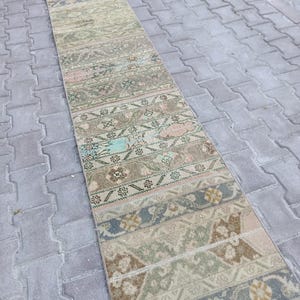May include: A long, narrow runner rug featuring a series of repeating rectangular panels. The design displays a muted colour palette of beige, sage green, dusty pink, and slate blue, with intricate floral and geometric patterns throughout the surface.