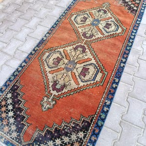May include: A rectangular, vintage-style rug with an orange field and geometric patterns. The rug features a central design with cream, blue, and purple accents. The border is dark blue with a repeating floral pattern. The rug is likely made of wool.