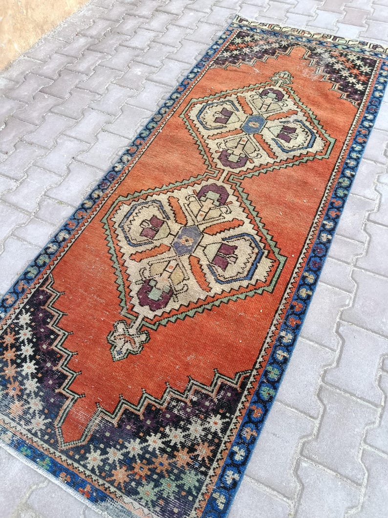 May include: A rectangular, vintage-style rug with an orange field and geometric patterns. The rug features a central design with cream, blue, and purple accents. The border is dark blue with a repeating floral pattern. The rug is likely made of wool.