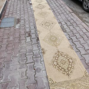 Pode incluir: Um tapete comprido e retangular bege com um padrão intrincado e repetitivo em tons de castanho e bege. O tapete é colocado sobre uma superfície de tijolo, mostrando o seu design e textura. O tapete é provavelmente para um corredor ou entrada.