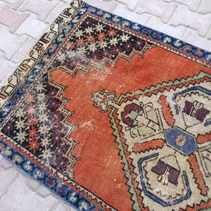 May include: An antique, rectangular rug with a distressed appearance. The rug features a central orange field with intricate geometric patterns in blue, purple, and beige. A blue border frames the design, adding a contrasting element to the overall aesthetic.