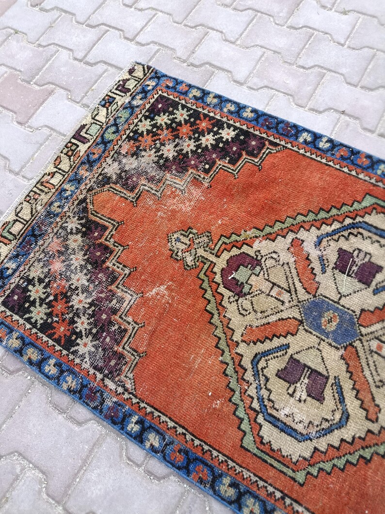 May include: An antique, rectangular rug with a distressed appearance. The rug features a central orange field with intricate geometric patterns in blue, purple, and beige. A blue border frames the design, adding a contrasting element to the overall aesthetic.