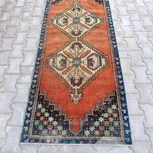 May include: A rectangular, vintage-style rug with an orange field and geometric patterns. The rug features a central diamond design in beige, blue, and burgundy, framed by a dark blue border with star-like motifs. The rug is placed on a gray brick surface.