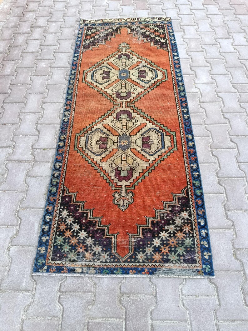 May include: A rectangular, vintage-style rug with an orange field and geometric patterns. The rug features a central diamond design in beige, blue, and burgundy, framed by a dark blue border with star-like motifs. The rug is placed on a gray brick surface.