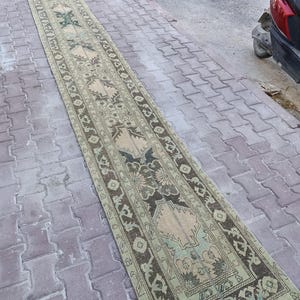 May include: A long, narrow runner rug featuring a faded, muted colour palette of beige, sage green, and dark brown. The rug displays a repeating geometric and floral pattern with intricate borders, finished with short, light-coloured fringe at the ends.