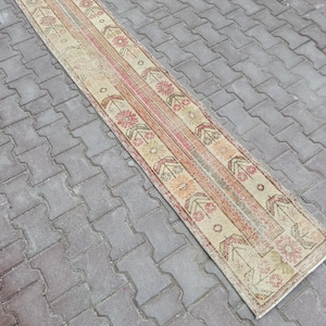 May include: A rectangular, vintage runner rug with a central stripe of pink and red hues, flanked by beige panels with floral and geometric patterns. The rug's design features a distressed, faded appearance.