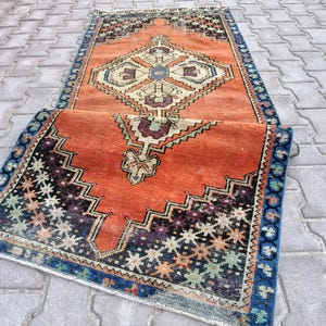 May include: An orange and blue vintage rug with a geometric pattern. The rug features a central diamond design in cream, blue, and purple, surrounded by a border of blue and black zigzags. The rug is partially rolled up, showcasing its intricate details.