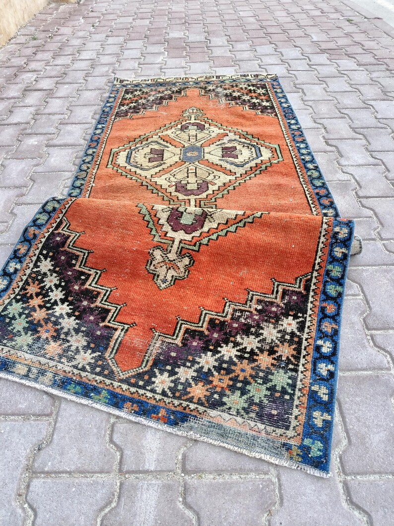 May include: An orange and blue vintage rug with a geometric pattern. The rug features a central diamond design in cream, blue, and purple, surrounded by a border of blue and black zigzags. The rug is partially rolled up, showcasing its intricate details.