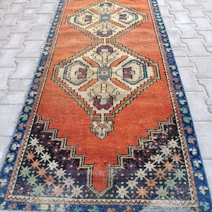 May include: An orange and blue vintage runner rug with geometric patterns. The rug features a central diamond design in beige, purple, and blue, framed by a border of blue and black geometric shapes. The rug's dimensions are approximately 3 x 8 feet.