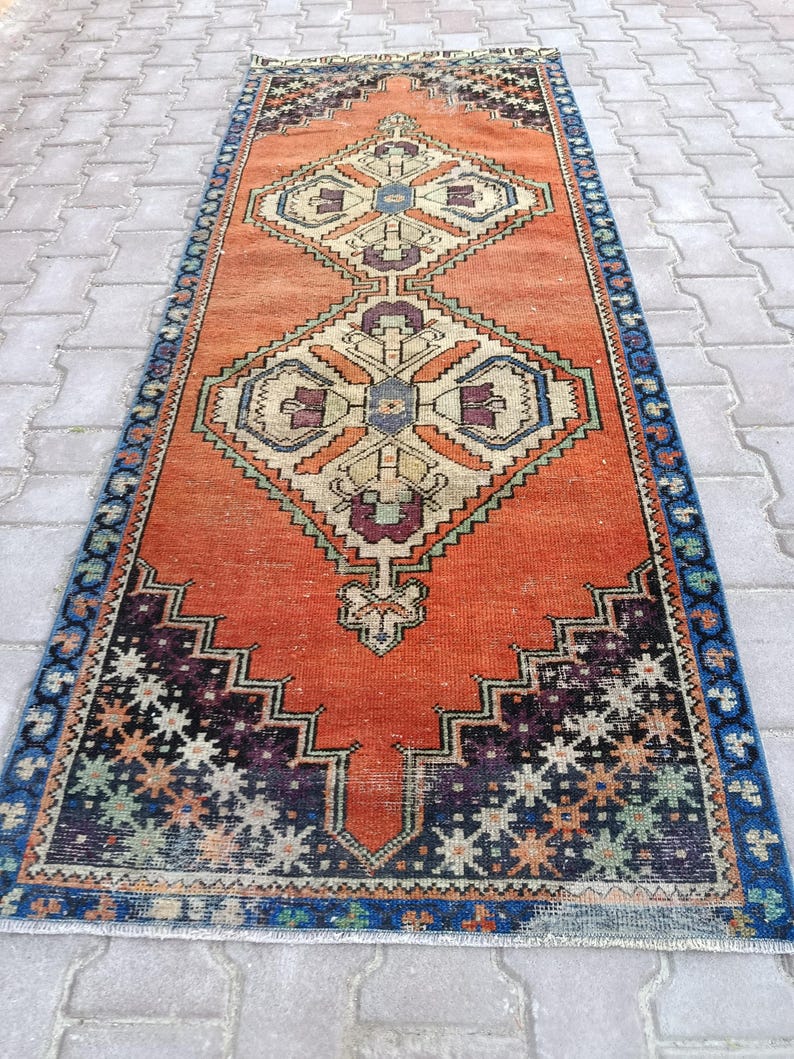May include: An orange and blue vintage runner rug with geometric patterns. The rug features a central diamond design in beige, purple, and blue, framed by a border of blue and black geometric shapes. The rug's dimensions are approximately 3 x 8 feet.