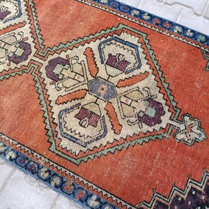 May include: An orange and blue vintage runner rug with geometric patterns. The rug features a central design with cream, purple, and orange details, framed by a blue border. The rug's design is symmetrical and detailed.