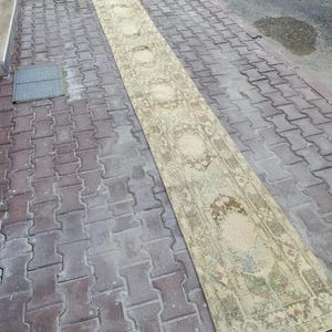 May include: A long, narrow rug with a faded floral design in beige, brown, and green. The rug is placed on a brick surface, highlighting its length and pattern. The rug's design features repeating floral motifs.