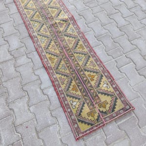 May include: A vintage runner rug with a geometric pattern. The rug features a central design in yellow, black, and pink, framed by a red border. The rug is rectangular and appears to be made of woven material.