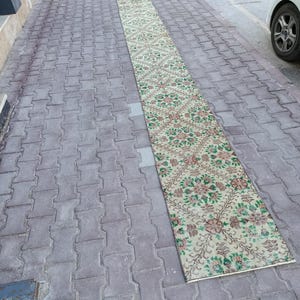 May include: A long, rectangular rug with a floral pattern in green, pink, and brown on a cream background. The rug is placed on a grey brick surface, highlighting its design and texture. The rug's length is emphasized in the outdoor setting.