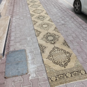 Könnte beinhalten: Ein langer, rechteckiger Teppich mit beigem Hintergrund und aufwendigen braunen Blumenmustern. Der Teppich liegt auf einer mit Ziegeln gepflasterten Oberfläche und zeigt seine Länge und sein Design. Die Ränder des Teppichs weisen einen detaillierten Rand auf.