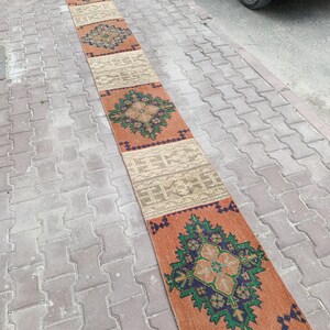 Könnte beinhalten: Ein langer, schmaler Läufer mit einem sich wiederholenden Muster aus abwechselnden rechteckigen Abschnitten. Das Design umfasst orangefarbene Felder mit dunkelblauen und grünen Blumenmedaillons sowie beige Felder mit geometrischen Linien.