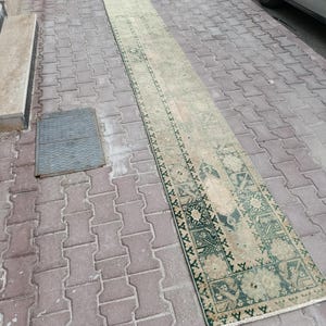 May include: A long, narrow, vintage rug with a faded, distressed appearance. The rug features a central field of beige with intricate green and dark green geometric and floral patterns along the borders and ends. The rug is placed on a brick surface.