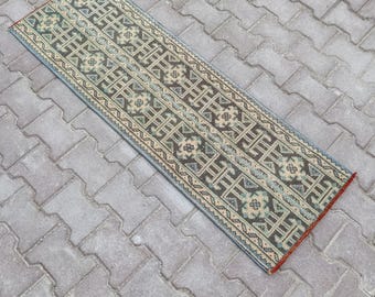 May include: A rectangular, patterned rug with a geometric design. The rug features a repeating pattern in shades of brown, beige, and blue. The edges are finished with a red border. The rug is placed on a gray brick surface.