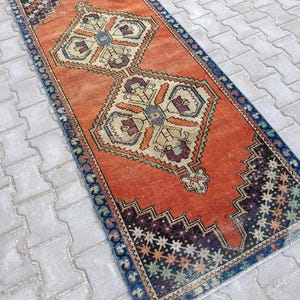 May include: An orange and blue vintage runner rug with a geometric pattern. The rug features a central design with beige, purple, and blue accents. The border is a dark blue with a repeating pattern. The rug is rectangular.
