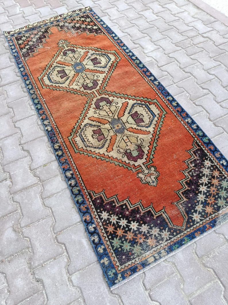 May include: An orange and blue vintage runner rug with a geometric pattern. The rug features a central design with beige, purple, and blue accents. The border is a dark blue with a repeating pattern. The rug is rectangular.