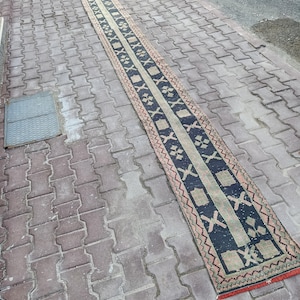 May include: A long, narrow rug with a central dark blue band featuring geometric patterns and a light beige border. The rug has a red fringe on the ends and is placed on a brick surface. The rug's design includes cross and diamond shapes.