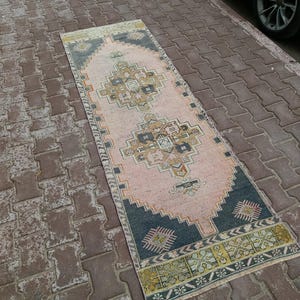 May include: A rectangular, vintage-style rug with a central pink field and intricate geometric patterns in shades of black, gold, and beige. The rug has a dark blue border and is placed on a brick surface.
