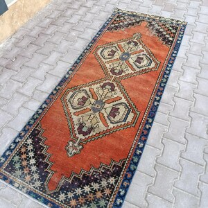 May include: An orange and blue vintage runner rug with geometric patterns. The rug features a central orange field with two large, symmetrical designs in beige, blue, and burgundy. The borders are dark blue with star-like patterns.
