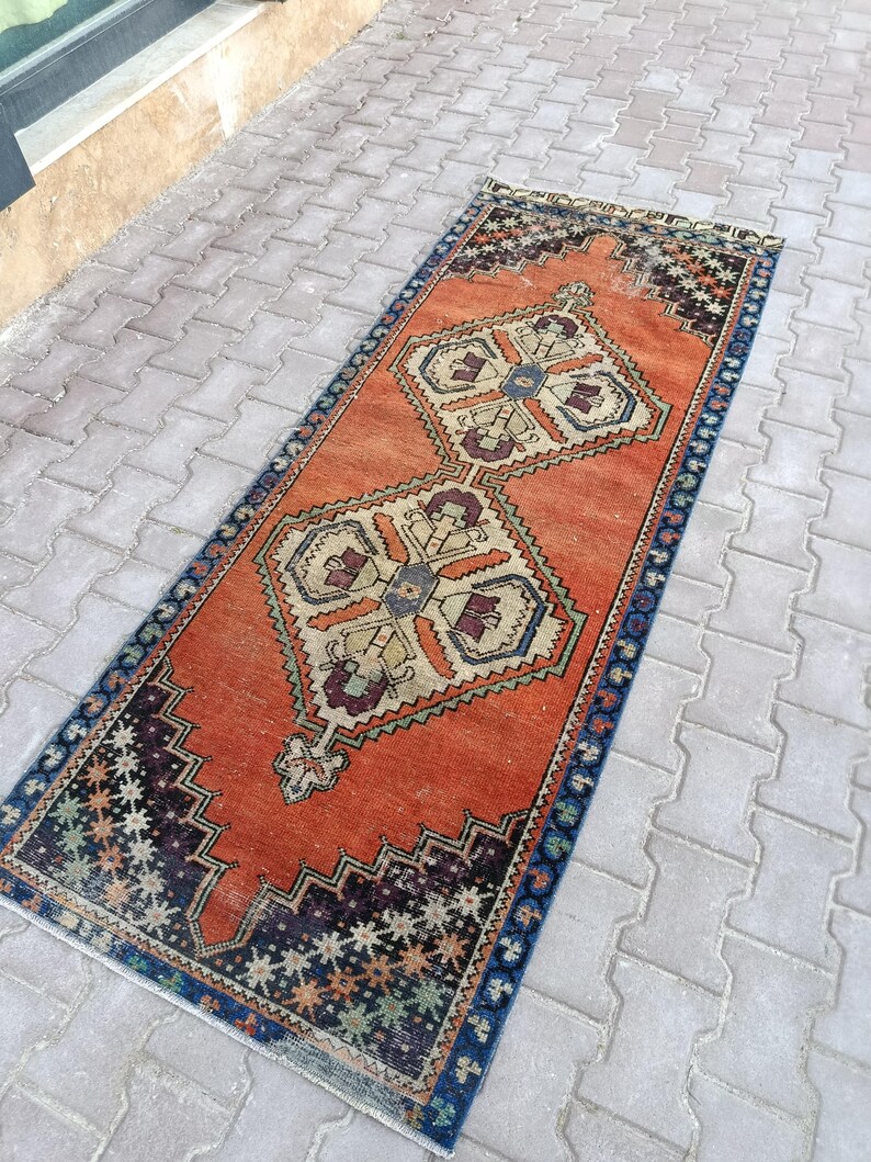 May include: An orange and blue vintage runner rug with geometric patterns. The rug features a central orange field with two large, symmetrical designs in beige, blue, and burgundy. The borders are dark blue with star-like patterns.