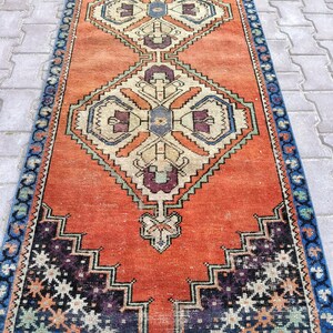 May include: An orange and blue runner rug with geometric patterns. The rug features a central orange field with diamond-shaped designs in cream, purple, and blue. The borders are blue with repeating floral motifs. The rug is likely made of wool.