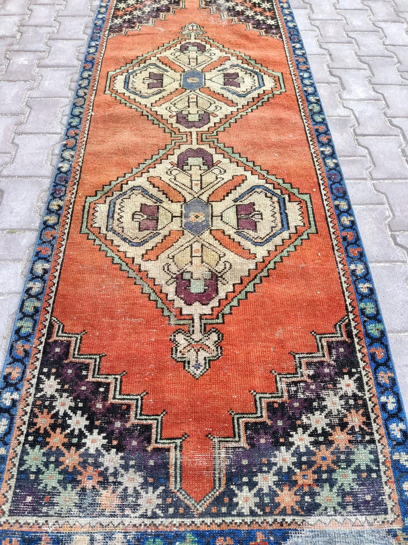 May include: An orange and blue runner rug with geometric patterns. The rug features a central orange field with diamond-shaped designs in cream, purple, and blue. The borders are blue with repeating floral motifs. The rug is likely made of wool.