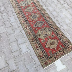 May include: A rectangular, red runner rug with an intricate geometric pattern. The rug features a central red field with repeating diamond and floral motifs in shades of beige, blue, and green. The border is dark blue with additional patterns.