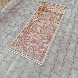 Könnte beinhalten: Ein rechteckiger, verblasster Teppich mit floralem Muster in Rot-, Beige- und Grüntönen. Der Teppich hat ein verwaschenes Aussehen und einen beigen Rand. Er liegt auf einer grauen Ziegeloberfläche.