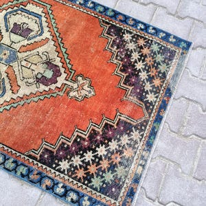 May include: A rectangular, vintage-style rug with a distressed appearance. The rug features a vibrant orange central field with intricate geometric patterns in cream, blue, purple, and black. A blue border frames the rug's edges.