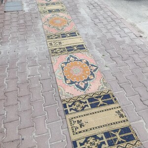 May include: A long, narrow runner rug featuring a repeating pattern of large, central floral medallions in shades of orange, pink, and dark blue. The design includes geometric borders in beige and navy blue, with intricate, symmetrical motifs throughout.