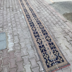Puede incluir: Una alfombra larga y estrecha con un patrón geométrico repetitivo en azul marino, beige y toques de rojo. La alfombra es rectangular y parece estar tejida a mano, con un diseño detallado y bordes con flecos. La alfombra está colocada sobre una superficie de ladrillo.