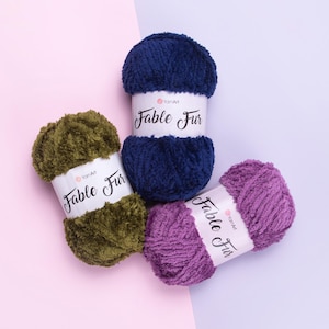 Op de afbeelding: Drie bollen YarnArt Fable Fur garen in marineblauw, olijfgroen en paars. Elke bol heeft een wit etiket met de tekst "Fable Fur". Het garen is zacht en pluizig, ideaal voor brei- of haakprojecten.