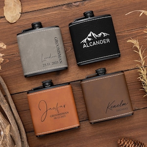 Puede incluir: Cuatro petacas personalizadas en cuero gris, negro, marrón y beige. Cada petaca tiene una tapa negra y costuras blancas. Las petacas están grabadas con nombres, fechas y la palabra "Groomsman" o un gráfico de montaña.