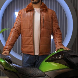 May include: A brown leather jacket with a fur collar and a zipper closure. The jacket has a quilted design and is worn over a white t-shirt and blue jeans. The person is standing next to a green motorcycle.
