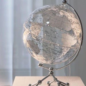 Puede incluir: Un globo terráqueo transparente con detalles geográficos, montado sobre un soporte de metal plateado. El interior del globo es visible, mostrando continentes y océanos. El soporte tiene una base de tres patas.