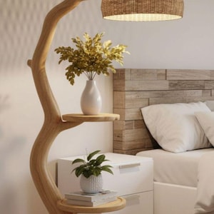 May include: A wooden floor lamp with a tree-like design. It features a woven lampshade and two round shelves. A white vase with greenery and a small potted plant are displayed on the shelves. The lamp is next to a white nightstand and bed.