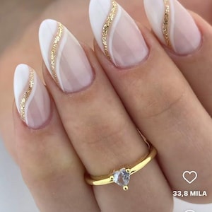May include: Oval-shaped nail design featuring a white and gold glitter accent. The nails have a nude base with a white curved design and a gold glitter stripe. A gold ring with a heart-shaped stone is also visible.