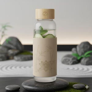 May include: A clear glass water bottle with a bamboo lid and a beige sleeve featuring a bamboo and lotus flower design. The bottle contains water, cucumber slices, and mint leaves. The bottle is sitting on a black stone.