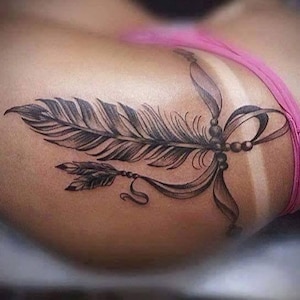Könnte beinhalten: Detailliertes Tattoo-Design mit schwarzer und grauer Feder. Das Tattoo zeigt eine große Feder mit komplizierten Details, eine kleinere Feder und eine dekorative Schleife mit Perlen. Das Design befindet sich auf einer Hautoberfläche, mit einem rosa Kleidungsstück sichtbar.