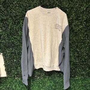 Könnte beinhalten: Ein graues Sweatshirt mit blau-weiß karierten Ärmeln. Das Sweatshirt hat eine kleine Grafik auf der Vorderseite und einen Rundhalsausschnitt. Die Ärmel sind lang und haben Knöpfe an den Manschetten.