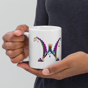 May include: White ceramic mug with a vibrant, artistic letter "W" design. The letter is decorated with green, purple, blue, and red elements, including hearts and dots. The mug is held in a person's hands.