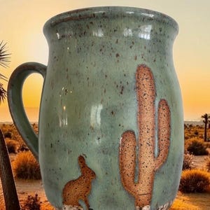 May include: A ceramic mug with a speckled sage green glaze featuring a textured, raised brown relief of a saguaro cactus and a small rabbit at the base. The mug has a curved handle and a smooth, glossy finish with a darker brown rim.