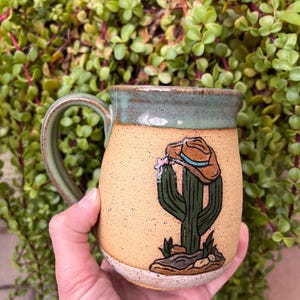 May include: A ceramic mug with a speckled tan body and a green glazed rim and handle. The front features a hand-painted illustration of a green saguaro cactus wearing a brown cowboy hat with a blue band, set on a rocky base.